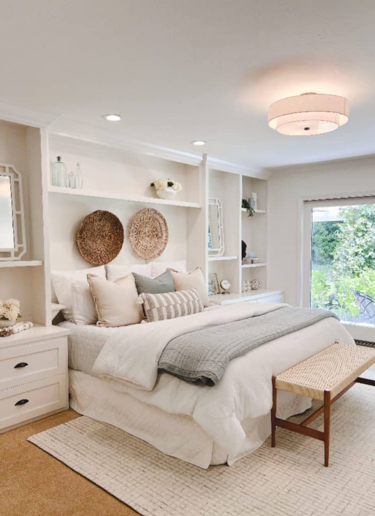 A bright, cozy bedroom with a neatly made bed, neutral bedding, decorative pillows, and woven baskets on the wall—perfect for relaxing with my favorite summer drinks while enjoying garden views from large windows. A wooden bench sits at the foot of the bed.
