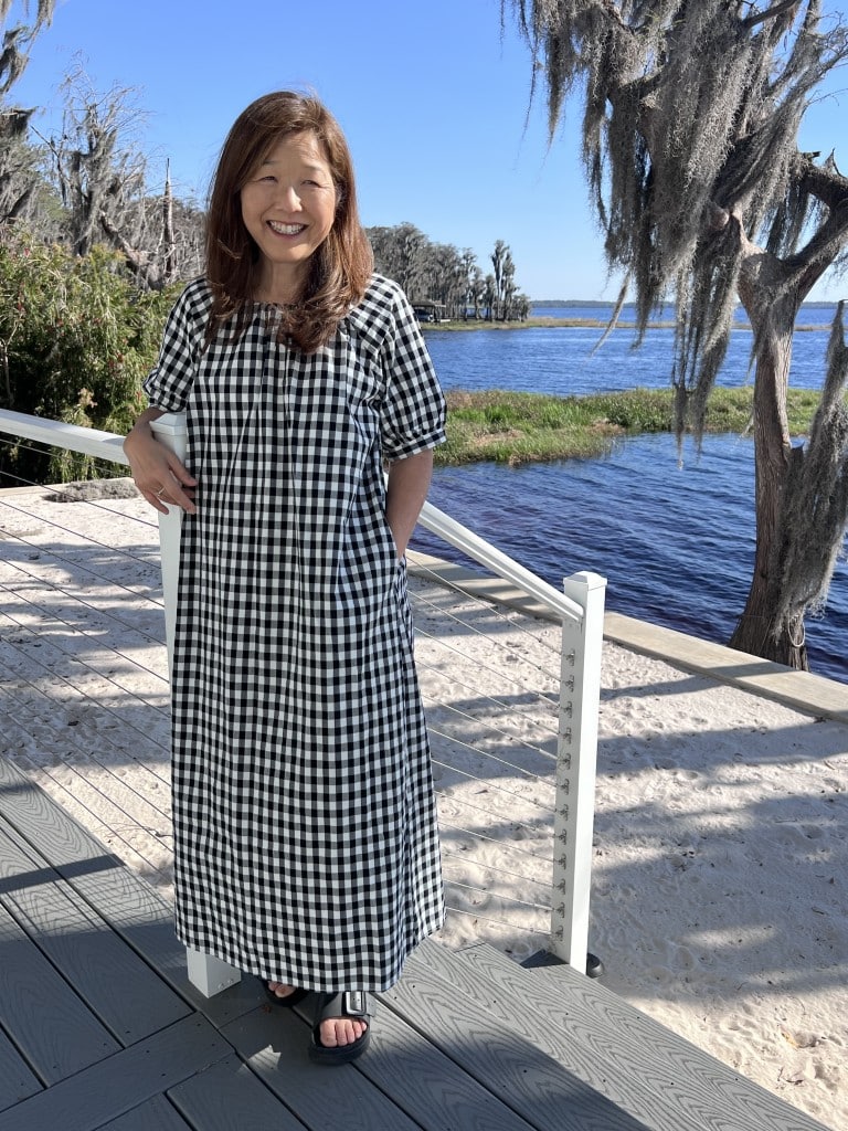 A woman with long brown hair smiles while standing on a sunlit deck by a lake, wearing one of the casual summer dresses for women—a black-and-white checkered dress and sandals. Moss-covered trees and blue sky create a serene backdrop.