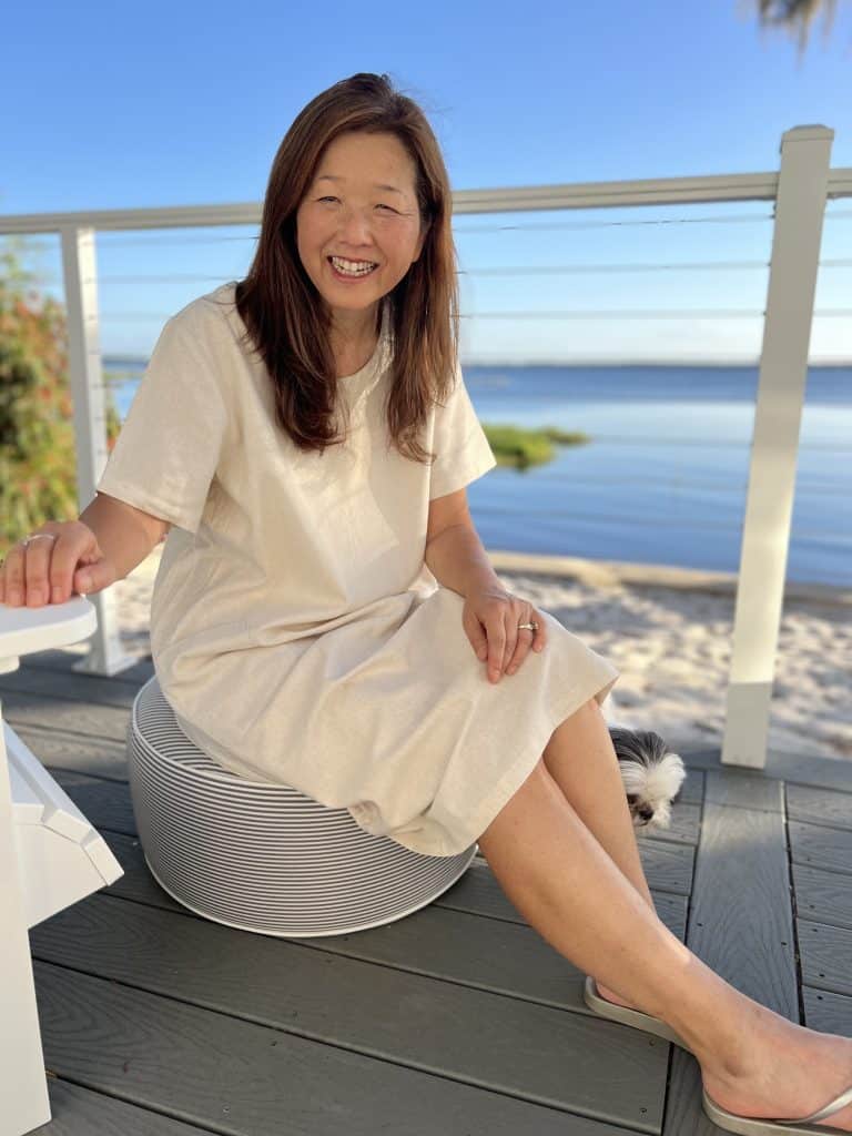 A woman in a cream-colored casual summer dress sits smiling on a striped round seat on a deck by the water, with greenery and a sandy area nearby. A small dog peeks out from behind her legs on the wooden floor.