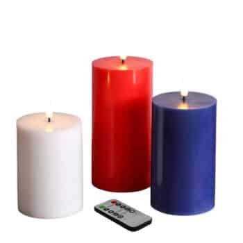 Three flameless LED candles in white, red, and blue, stand upright next to each other. A black and white remote control with colorful buttons is placed in front of the candles.