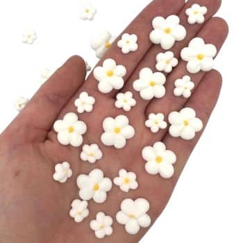 A hand holding several small, white, five-petal flower decorations with yellow centers—reminiscent of my favorite floral desserts—with a few more scattered nearby.
