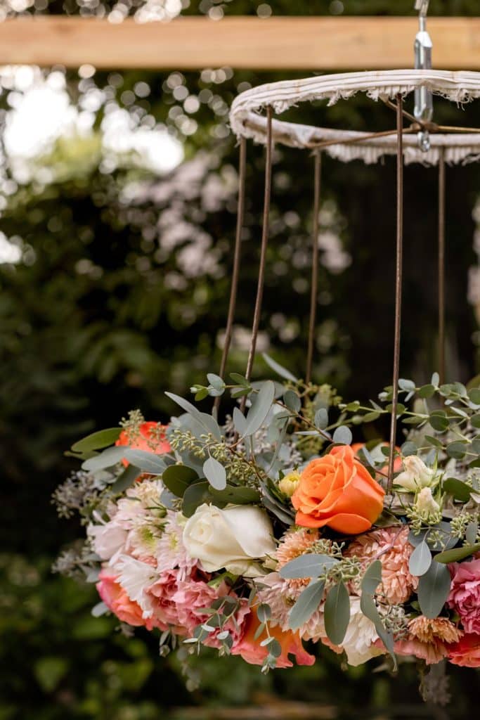 A hanging floral arrangement shaped like a chandelier, decorated with orange, white, and pink roses, greenery, and suspended outdoors against trees—my favorite summer decor to make for a stunning seasonal display.
