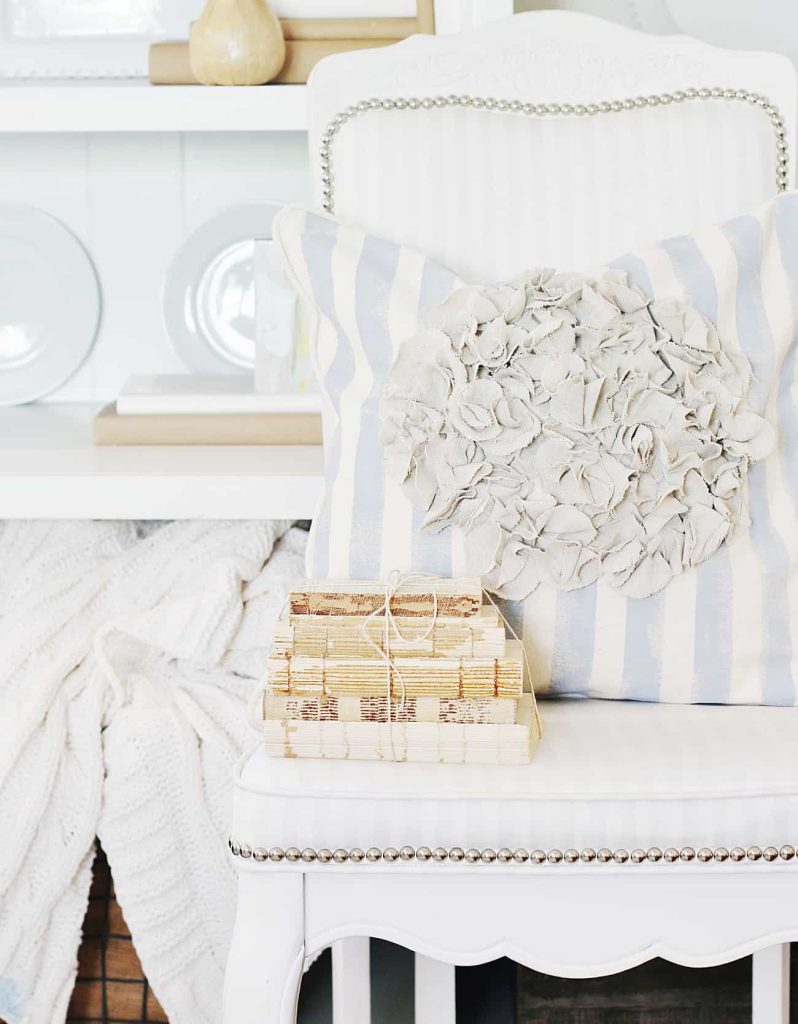 A white chair with a blue and white striped pillow featuring fabric flowers, one of my favorite summer decor to make. On the chair sits a stack of tied vintage books, with a white shelf, plates, and a folded cream blanket in the background.