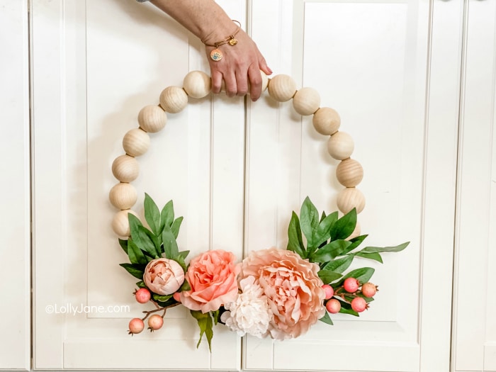 A hand holds a round wreath made of large wooden beads, decorated at the bottom with pink and white artificial flowers, green leaves, and small pink berries—my favorite summer decor to make. The background is a white cabinet.