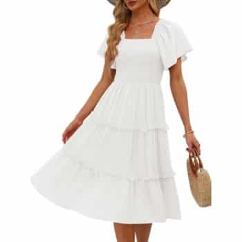A woman wearing a white, short-sleeve, tiered midi dress and a wide-brimmed hat holds a round woven handbag. Her wavy hair is loose, and she is posing with one hand holding her dress.