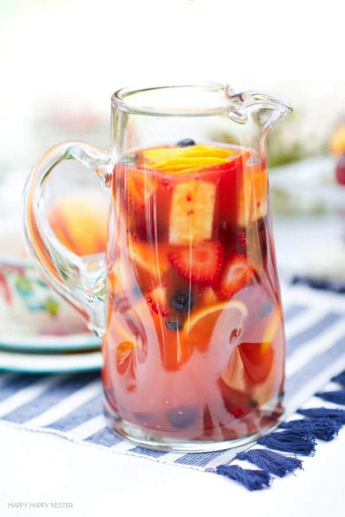 A clear glass pitcher filled with fruit punch—one of my favorite summer drinks—contains visible slices of oranges, strawberries, and whole blueberries, and sits on a blue and white striped cloth.