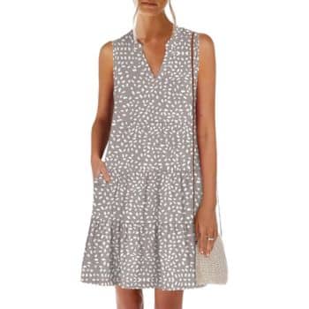 A woman wearing a sleeveless, gray dress with a white abstract dot pattern and a V-neckline, holding a woven shoulder bag, standing against a plain background.