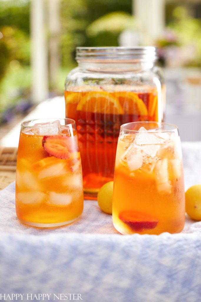 A glass jar and two glasses filled with iced tea, sliced strawberries, and other fruit—one of my favorite summer drinks—sit on a cloth outdoors against a bright, blurred garden backdrop.