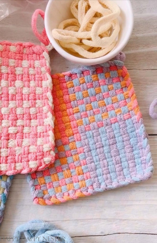 Two colorful woven potholders—my favorite summer decor to make—one in pink and white, the other in pastel blue, orange, purple, and pink, rest on a wooden surface beside a white bowl filled with unused loops.