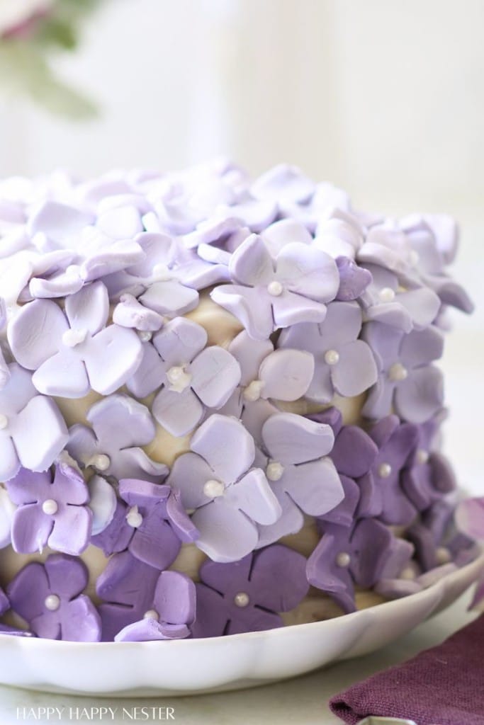 A cake decorated with layers of purple and lavender fondant flowers, each with a small white center, sits on a white plate. The lovely floral design makes it one of my favorite floral desserts.