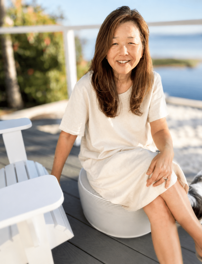 A woman with long brown hair, wearing a light-colored dress, sits smiling on a round white seat on a sunny outdoor deck near water, dreaming about my favorite summer drinks, with a white chair and greenery in the background.
