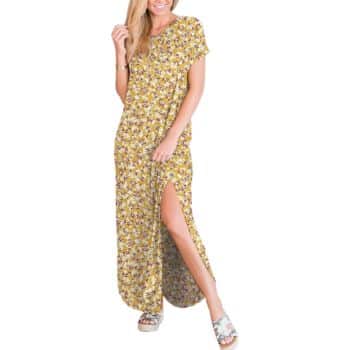 A woman wearing a yellow floral short-sleeve maxi dress with a thigh-high slit, posing with one hand in her hair and the other holding the dress. She is wearing platform sandals.