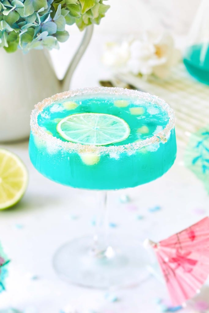 A blue cocktail in a glass with a sugared rim, garnished with lime and ice cubes, sits on a white surface with pastel decorations, a pink drink umbrella, and a cut lime—one of my favorite summer drinks for any sunny day.