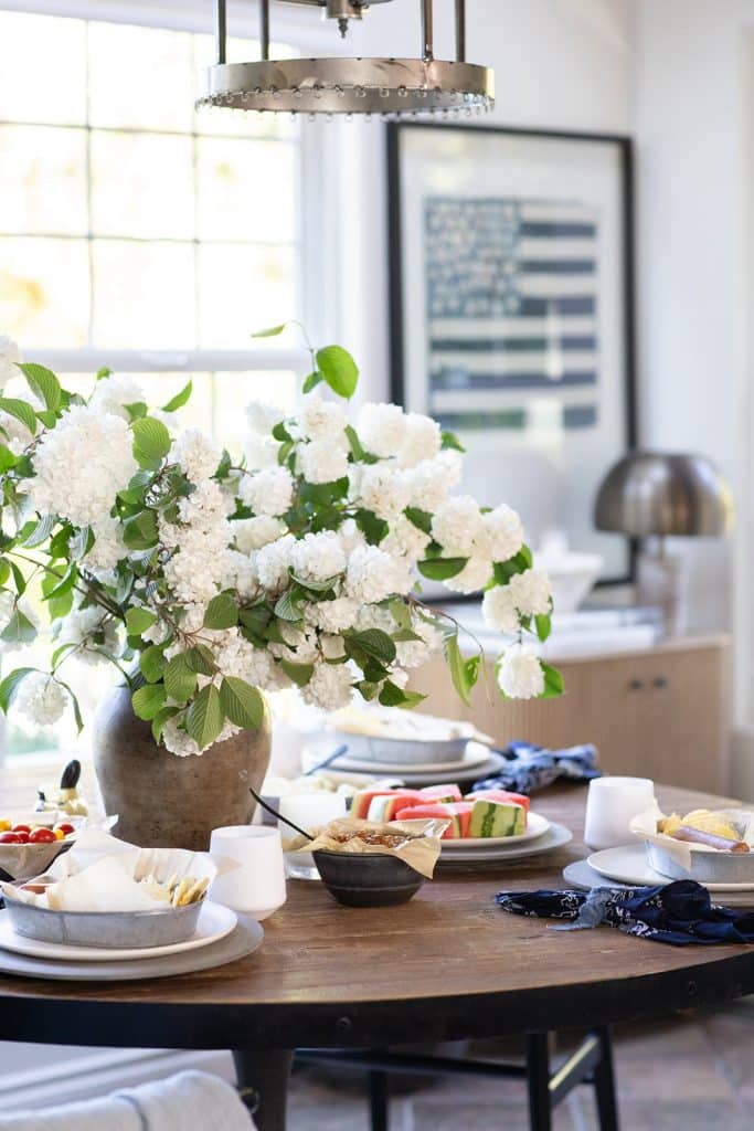 A round wooden table set for a meal with plates of fruit, cheese, and bread, a large vase of white flowers as the centerpiece, and a patriotic artwork on the wall in a bright, sunlit dining room.