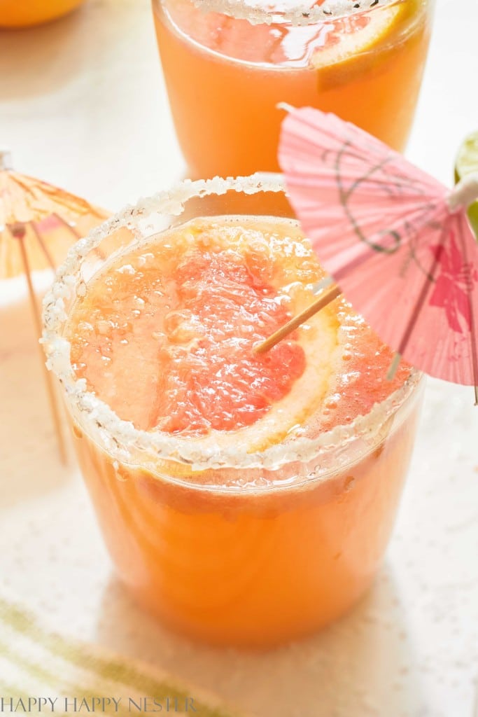 A close-up of a grapefruit cocktail in a glass with a salted rim, garnished with a grapefruit slice and a pink paper umbrella—one of my favorite summer drinks. Another similar drink is partially visible in the background.