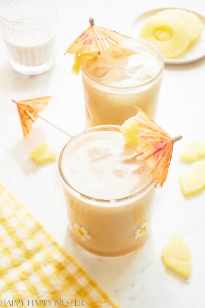 Two glasses of a creamy drink with ice, garnished with pineapple chunks and paper umbrellas, sit on a white surface—my favorite summer drinks—next to a yellow checkered cloth and a plate with sliced pineapple in the background.