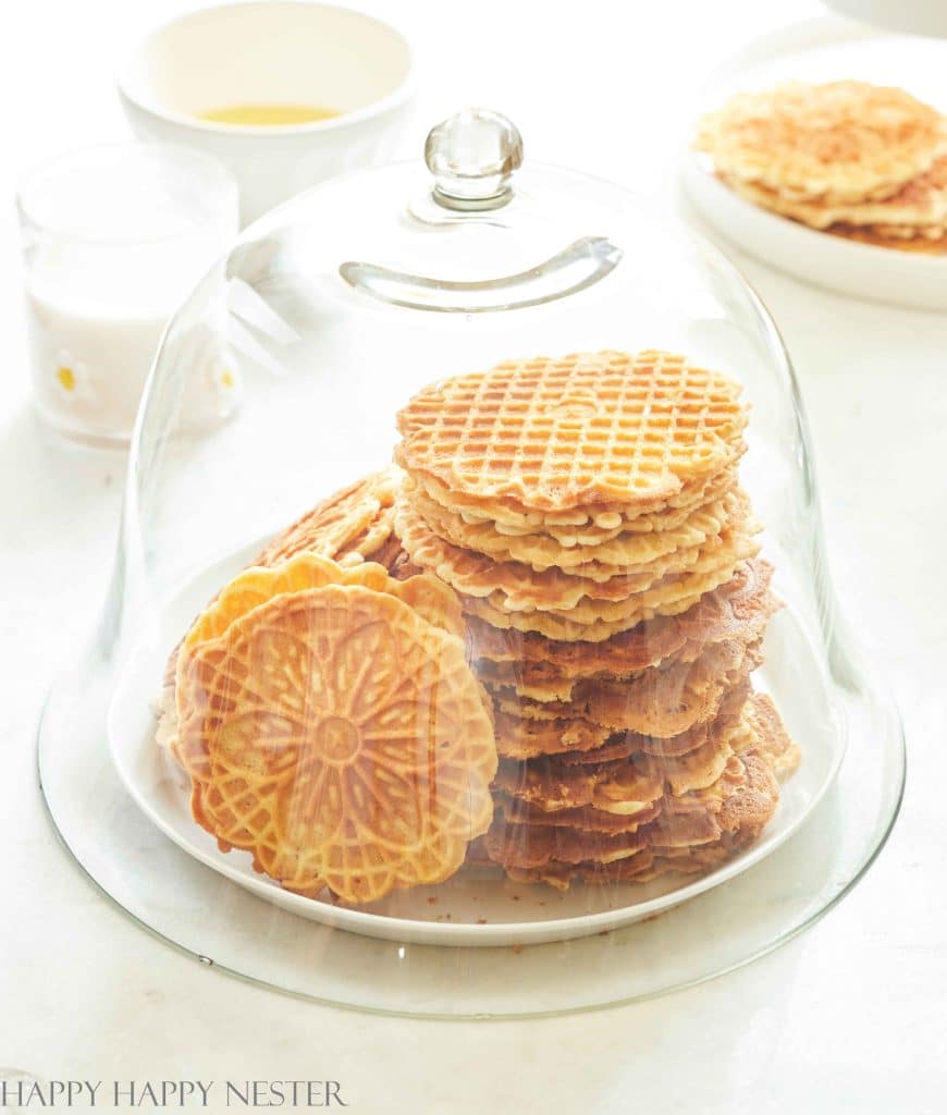 A stack of thin, round pizzelle cookies—one of my favorite floral desserts—is displayed on a white plate under a glass dome. More cookies, a cup of milk, and a bowl are visible in the background on a bright white surface.