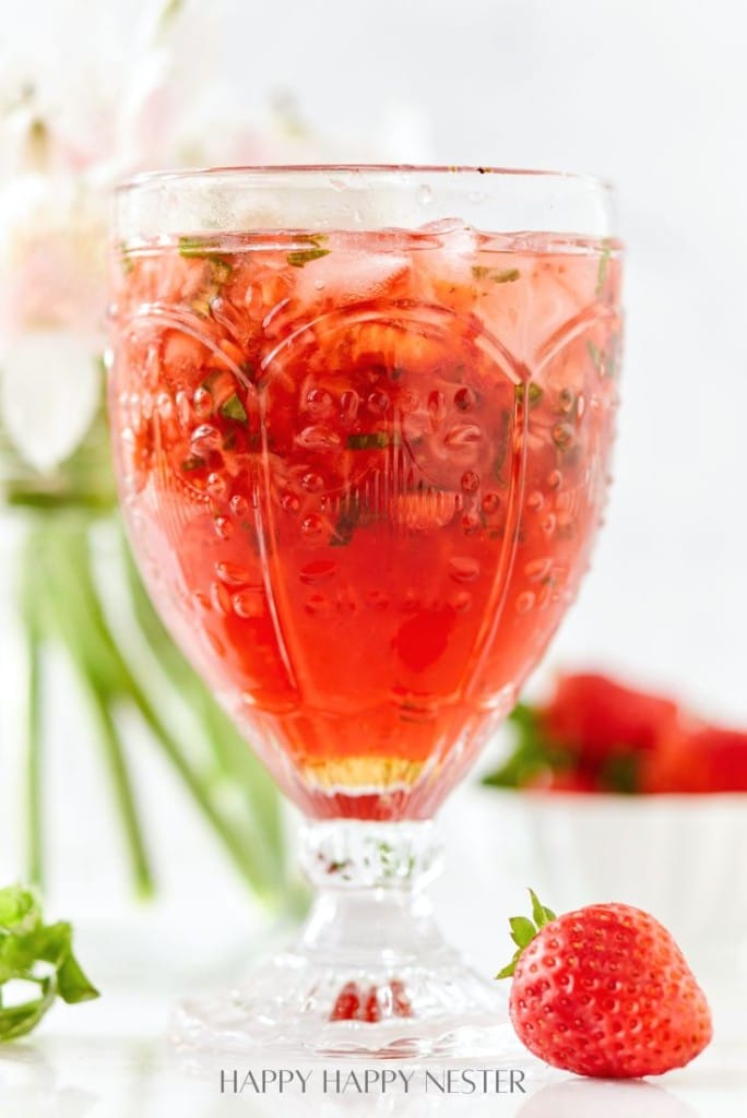 A glass filled with a red strawberry drink, ice, and mint, garnished with strawberry pieces. This refreshing treat is one of my favorite summer drinks, perfect alongside fresh strawberries and blooms blurred in the background.