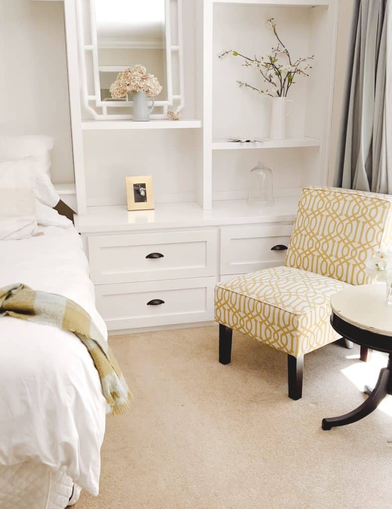 A cozy bedroom corner with a bed, white built-in shelves and drawers, a yellow patterned chair, and a round black table with flowers—featuring my favorite summer decor to make, like a framed photo and vase with branches for a fresh touch.