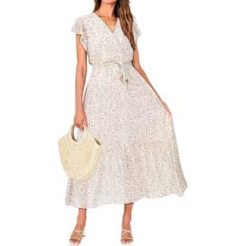 A woman wearing a light-colored, floral-patterned maxi dress with short flutter sleeves and a tie waist, holding a woven handbag and wearing strappy sandals.