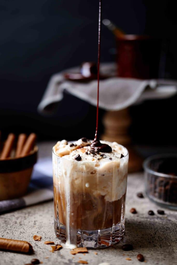 A glass of iced coffee, one of my favorite summer drinks, is topped with whipped cream and chocolate shavings, while chocolate syrup is drizzled on top. Coffee beans and cinnamon sticks are scattered nearby on the surface.