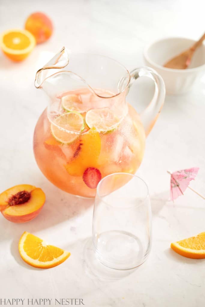 A glass pitcher filled with one of my favorite summer drinks—a peach and citrus blend with ice, peach slices, orange slices, and lime—sits on a white surface with a clear glass, fruit slices, and a pink cocktail umbrella nearby.