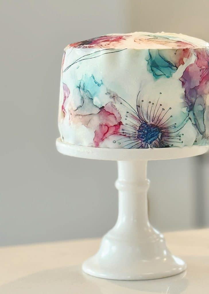 A round cake on a white stand, decorated with watercolor-style pastel flowers in shades of pink, blue, and purple, with delicate black line accents—one of my favorite cakes for its charming and artistic design.