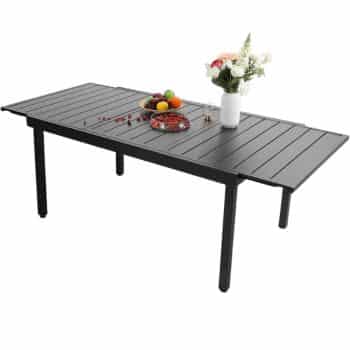 A black rectangular dining table with a slatted surface, set with a vase of flowers, a bowl of fruit, and a plate of assorted snacks on top.