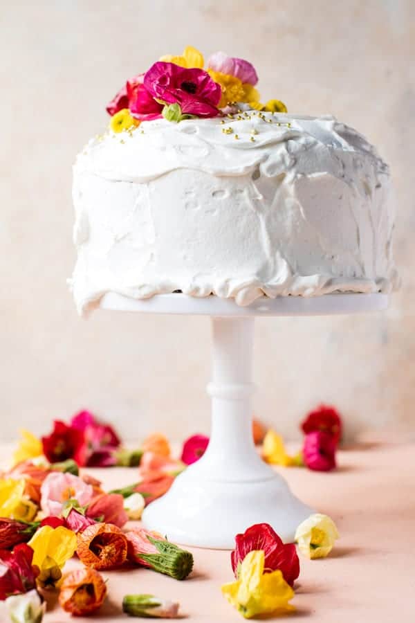 A white frosted cake, one of my favorite cakes, decorated with colorful edible flowers on top and displayed on a white cake stand. More flowers are scattered around the base on a light peach surface.