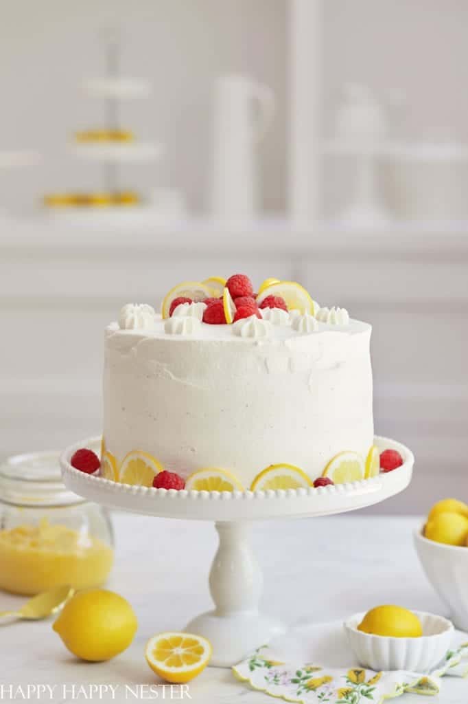 A white frosted cake—one of my favorite cakes—decorated with raspberries and lemon slices sits on a white cake stand. More lemons, raspberries, and a jar of lemon curd are on the table in the bright, airy setting.