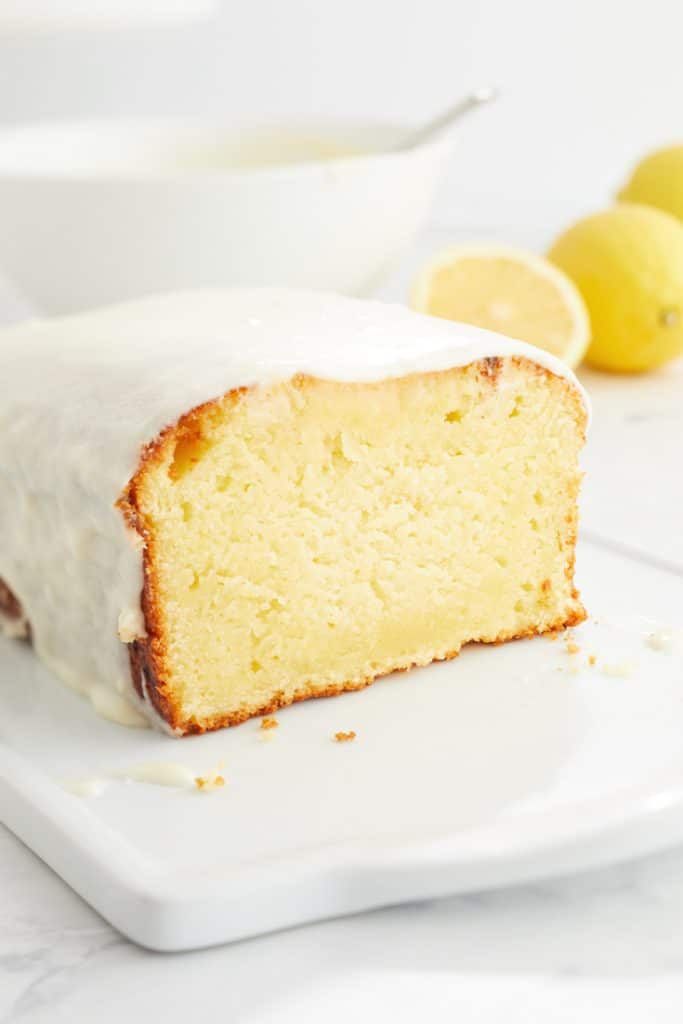 A loaf of lemon cake—one of my favorite cakes—with glossy icing, sliced to reveal a moist, yellow interior. Lemons and a bowl are blurred in the background on a white surface.