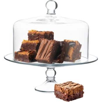 Glass cake stand with a dome lid displaying a stack of chocolate brownies—one of my favorite cakes. One brownie sits outside the stand on the surface below.