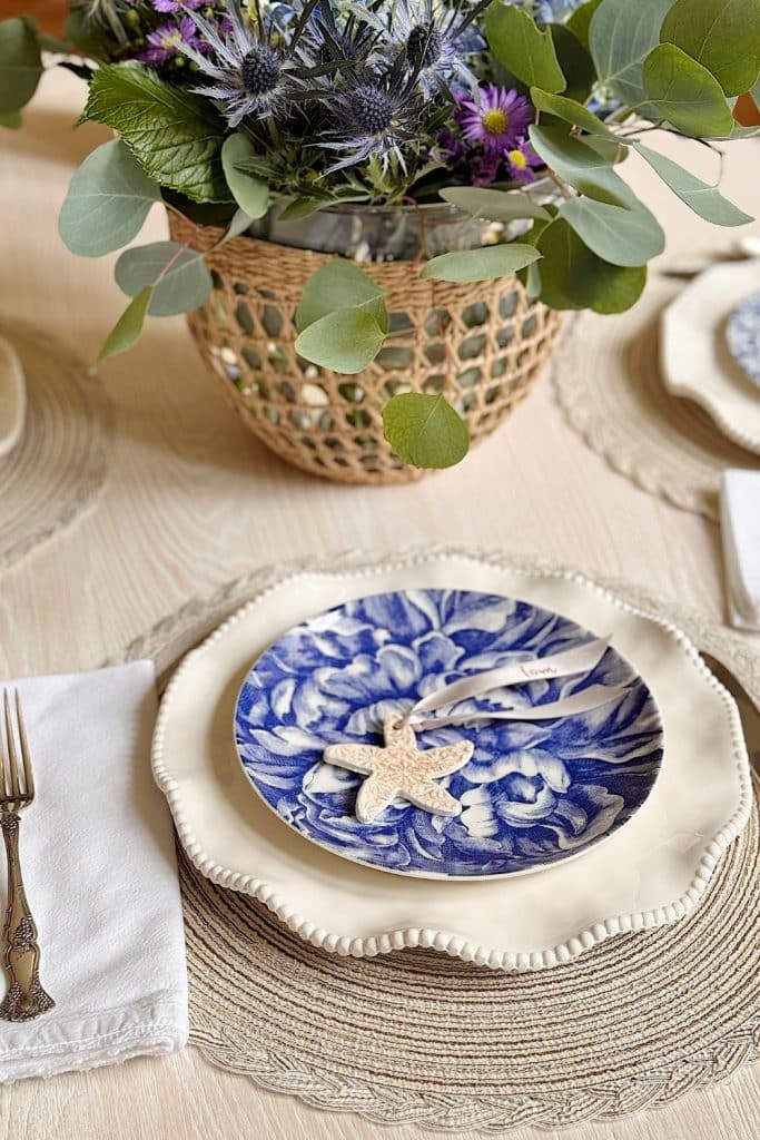 A table setting with a white napkin, silver fork, cream-colored plate, blue floral-patterned plate, and a starfish-shaped decoration. A basket of purple flowers and greenery is in the background.