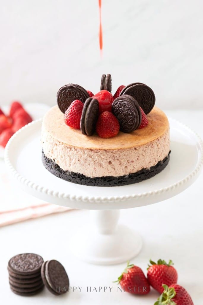 A cheesecake with an Oreo cookie crust, topped with whole strawberries, Oreo cookies, and cherries, sits on a white pedestal stand—one of my favorite cakes. Fresh strawberries and Oreos surround it as a drizzle is poured from above.