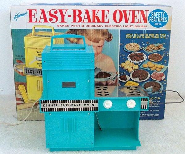 A vintage blue Easy-Bake Oven toy sits in front of its original box, which shows a young girl baking her favorite cakes and displays pictures of various pretend baked goods on plates.