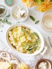 A dish of mashed potatoes topped with butter and fresh herbs sits on a table, surrounded by a glass of drink, a plate of butter, garlic cloves, a jar of herbs, and yellow flowers.