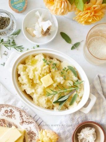 A dish of mashed potatoes topped with butter and fresh herbs sits on a table, surrounded by a glass of drink, a plate of butter, garlic cloves, a jar of herbs, and yellow flowers.