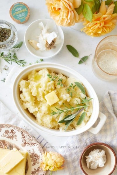 A dish of mashed potatoes topped with butter and fresh herbs sits on a table, surrounded by a glass of drink, a plate of butter, garlic cloves, a jar of herbs, and yellow flowers.