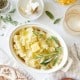 A dish of mashed potatoes topped with butter and fresh herbs sits on a table, surrounded by a glass of drink, a plate of butter, garlic cloves, a jar of herbs, and yellow flowers.