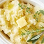 A close-up of creamy Instant Pot mashed potatoes in a white dish, topped with a pat of melting butter and garnished with fresh sprigs of rosemary and sage.