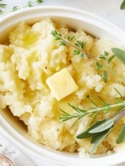 A close-up of creamy Instant Pot mashed potatoes in a white dish, topped with a pat of melting butter and garnished with fresh sprigs of rosemary and sage.