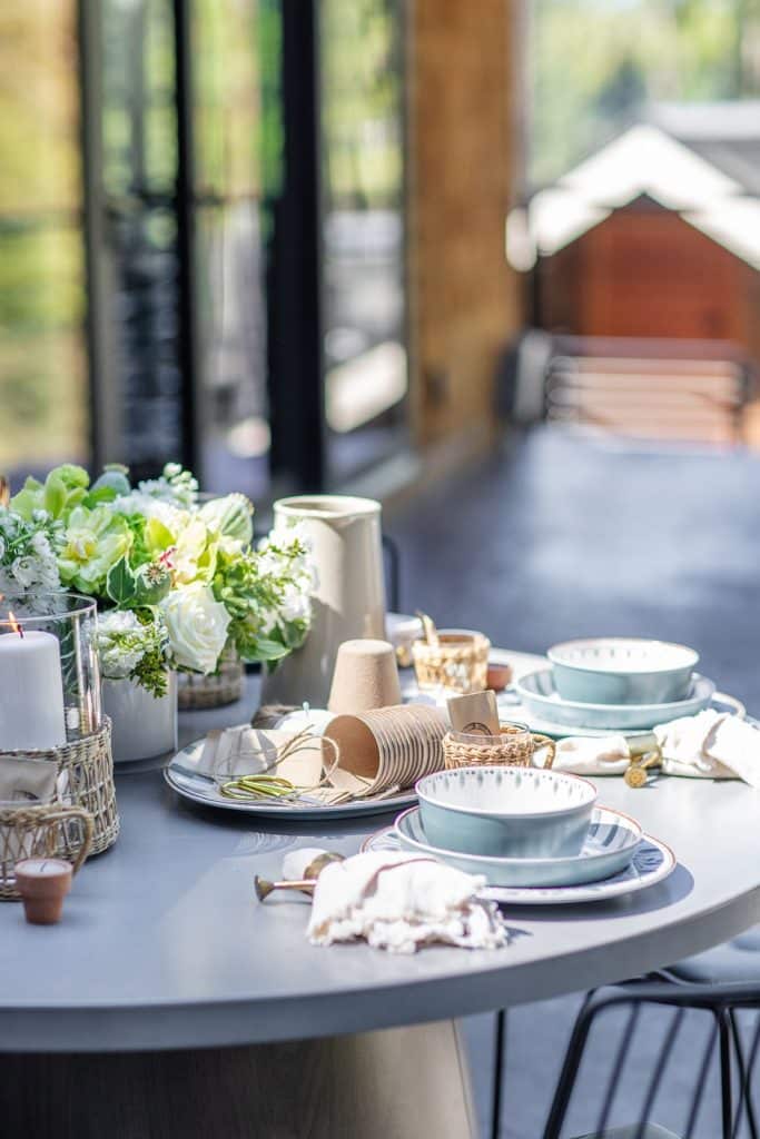 A round table set for a meal with blue and white dishes, napkins, cutlery, a pitcher, candles, and a bouquet of white and green flowers. Sunlight fills the modern indoor space in the background.