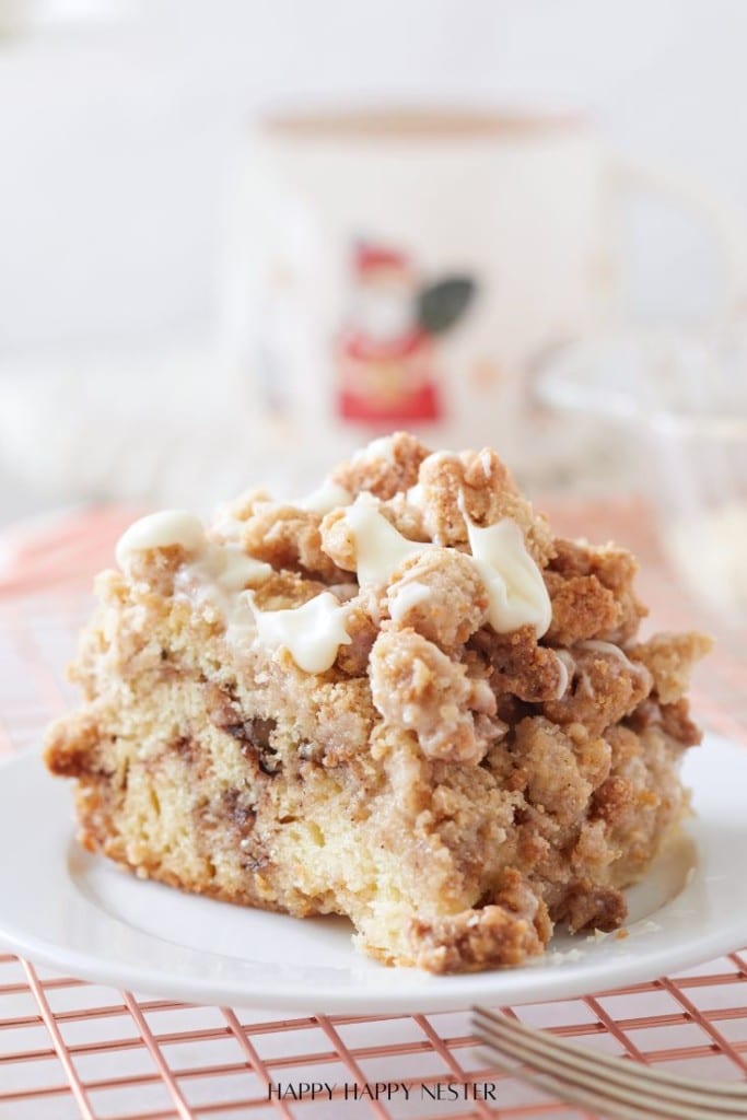 A slice of crumb-topped coffee cake—one of my favorite cakes—with cinnamon swirls and white icing sits on a white plate, set on a pink wire rack, with a blurred mug in the background.