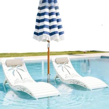Two white lounge chairs with palm tree cutouts sit partially submerged in a pool beneath a blue and white striped umbrella, complemented by outdoor tables and chairs in the grassy poolside area.
