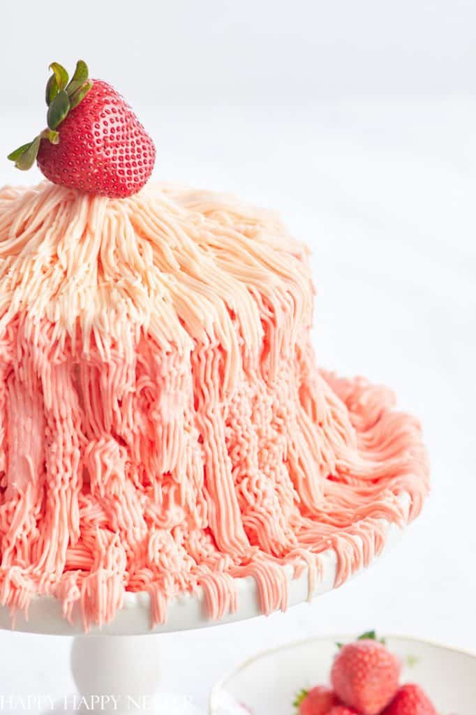 A pink and cream ombre cake with textured, wavy frosting—one of my favorite cakes—is topped with a fresh strawberry and sits on a white cake stand. A plate with strawberries is visible beside the cake.