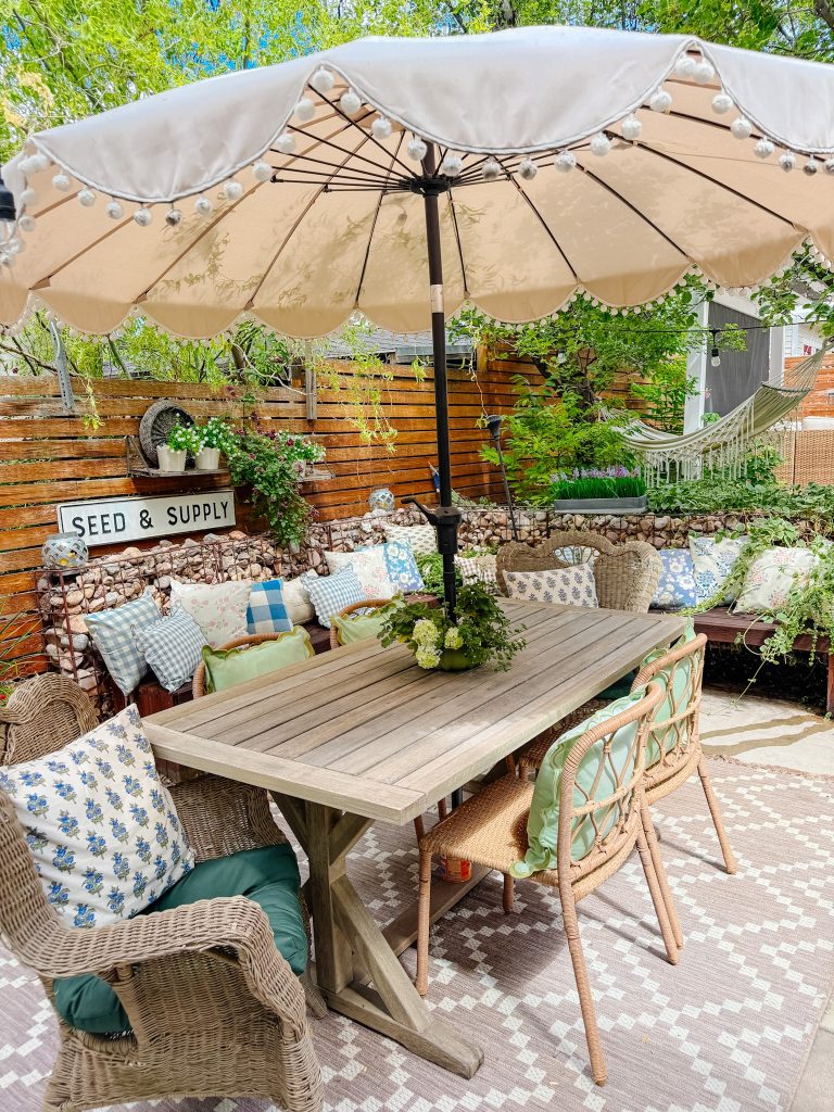 A cozy outdoor patio with a wooden table, wicker chairs, and bench seating covered in patterned pillows. A large cream umbrella shades the area, surrounded by greenery and a "Seed & Supply" sign on the wooden fence.