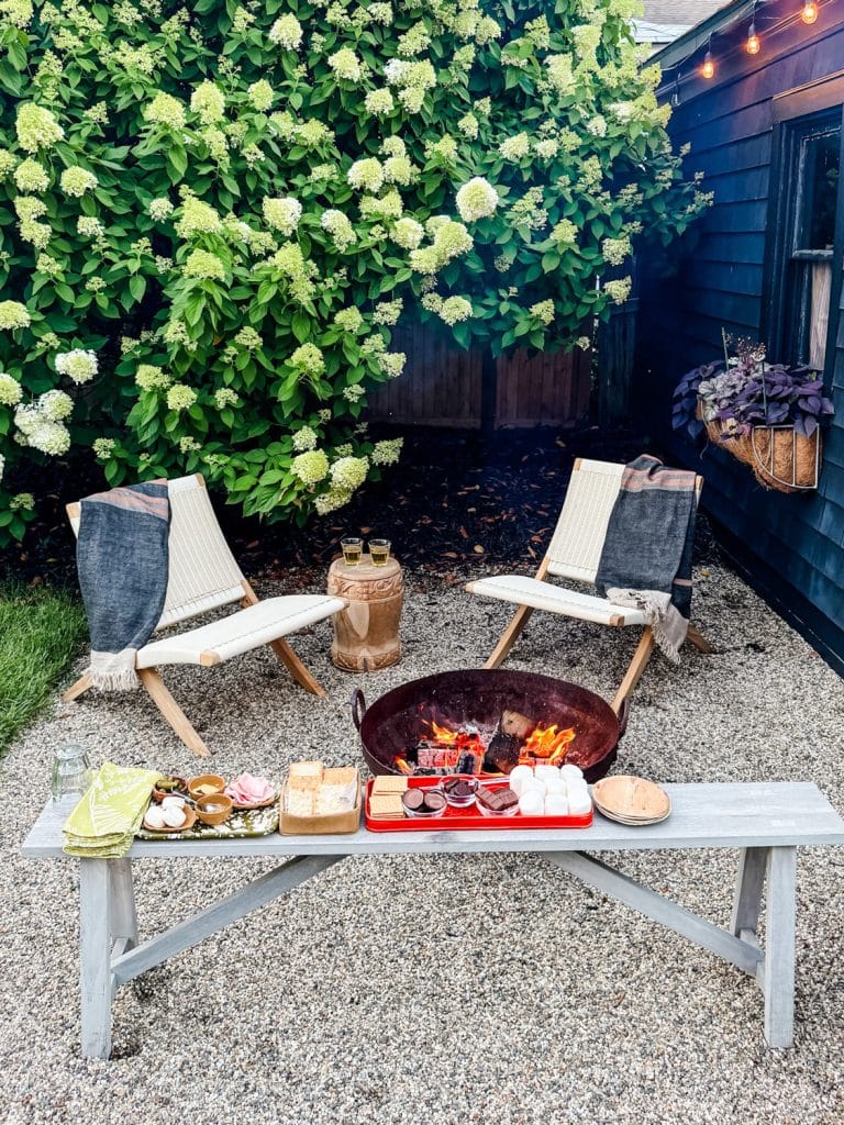 Two chairs with blankets sit around a fire pit in a gravel yard, with a bench holding s'mores ingredients in the foreground. Lush green hydrangea bushes and string lights provide a cozy outdoor setting.
