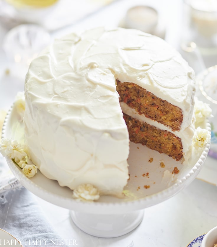 A two-layer carrot cake—one of my favorite cakes—covered in creamy white frosting sits on a white cake stand, with a large slice cut out to reveal the moist, textured interior. Small white flowers decorate the base of the cake.