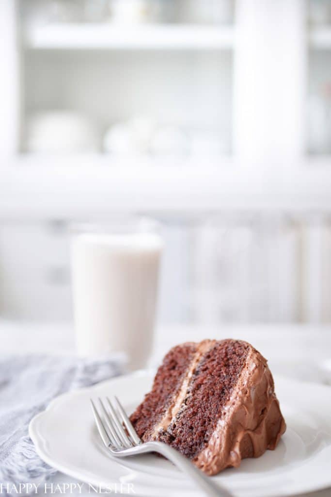 A slice of chocolate cake with chocolate frosting, one of my favorite cakes, sits on a white plate with a fork, next to a glass of milk, on a light-colored table. The background is softly blurred.