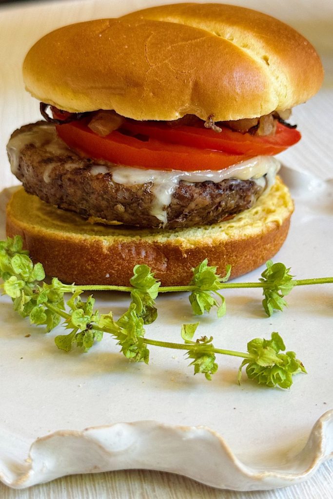 A cheeseburger with tomato slices and grilled onions on a bun sits on a white plate, inspired by the flying dutchman burger recipe, with a sprig of fresh green herbs beside it.